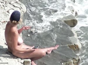 Курящая голая блондинка с телефоном сидит на берегу моря